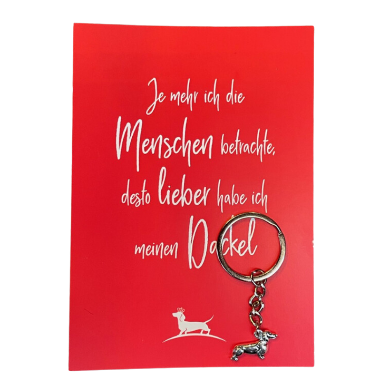 Rote Spruchkarte mit weißer Schrift, die sagt: „Je mehr ich die Menschen betrachte, desto lieber habe ich meinen Dackel“, begleitet von einem silbernen Schlüsselanhänger in Form eines Dackels.